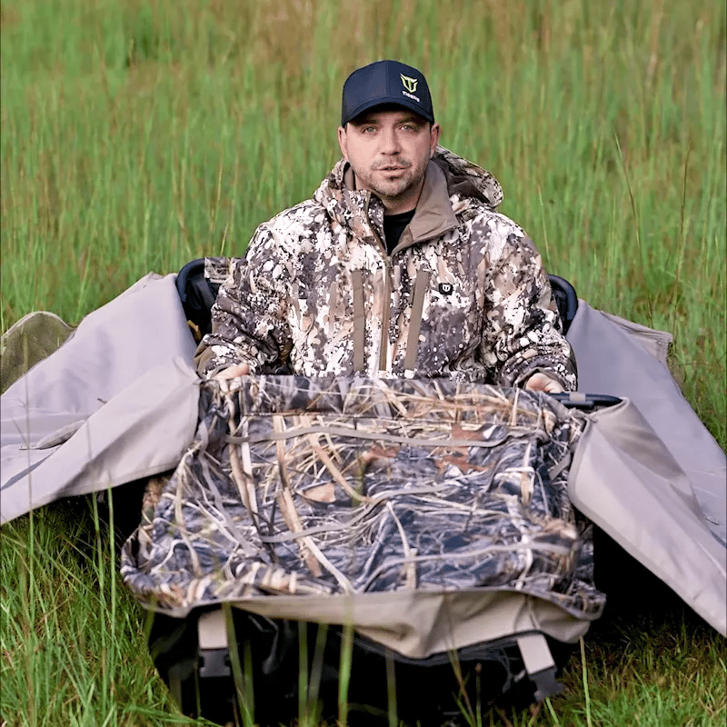 A hunter is Tidewe Layout Blind with Adjustable Height Duck Hunting Blind: A man sitting in a chair in a field, wearing a hat, holding a camouflage bag.