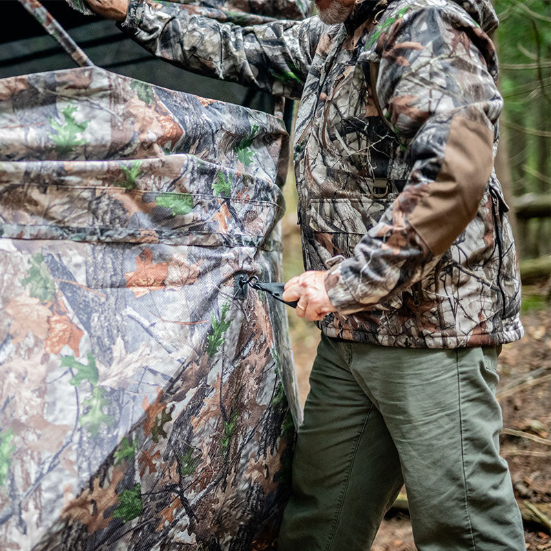 A man in camouflage outfit fixing a tent, a person's legs in green pants, and a person in a camouflage jacket, showcasing TideWe See Through Hunting Blind for 3-4 hunters.