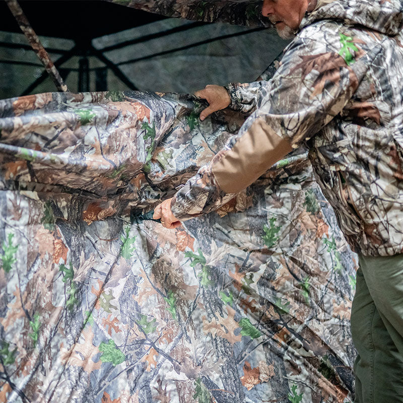 A man in camouflage clothing holds a TideWe See Through Hunting Blind, featuring one-way see-through panels for a wide view angle. Accommodates 3-4 hunters, noise-free windows, and durable construction.