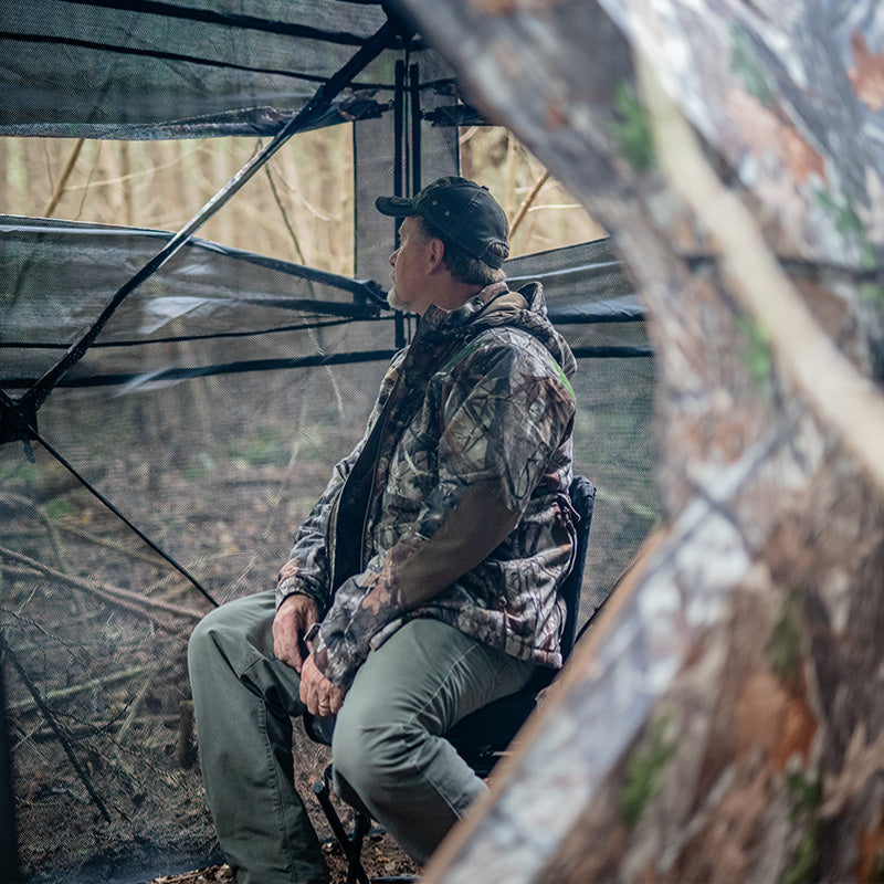 A man sits in a TideWe See Through Hunting Blind in a forest. 3-4 person pop-up ground blind with noise-free windows for undetected hunting. Dimensions: 80 height, 88.6 hub to hub.
